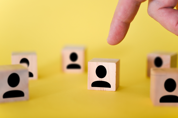 A finger poised above a wooden block printed with a person icon, surrounded by similar blocks on a yellow background, symbolising selecting a candidate.