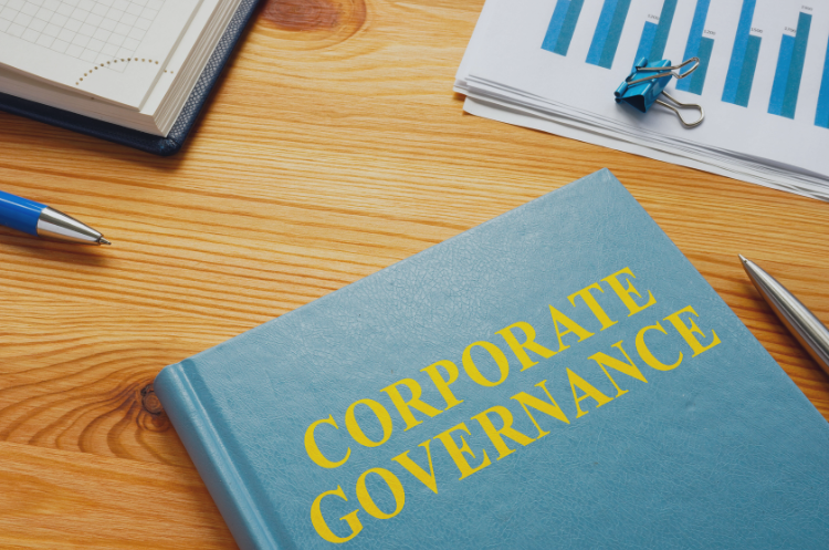 A blue book with the title "Corporate Governance" written in large yellow letters lies on a wooden desk surrounded by office items, including charts, documents, a pen, and a notebook.