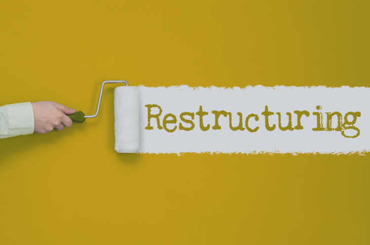 A hand using a paint roller to apply a stripe of white paint on a yellow wall, revealing the word "Restructuring" to symbolize corporate transformation and renewal.