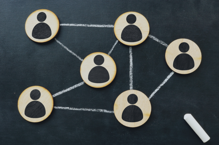 Wooden discs with person icons are arranged on a chalkboard and linked together with a network of hand-drawn chalk lines, symbolizing team connectivity and organizational alignment.