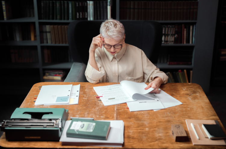 A senior Chief Knowledge Officer with glasses sitting at a desk in a library, carefully reviewing documents to manage and evaluate organizational knowledge.