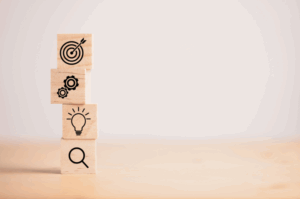 A stack of four wooden blocks on a light background, each displaying a key business strategy icon: a magnifying glass for research, a lightbulb for ideas, gears for execution, and a bullseye target for goals.
