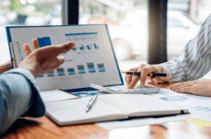 Operations analysts reviewing financial reports and performance charts during a strategic business meeting. One analyst points at a graph on a clipboard while the other uses a calculator to discuss data-driven insights and business strategy planning.