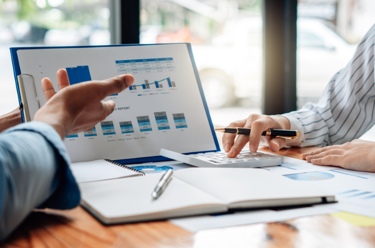 Operations analysts reviewing financial reports and performance charts during a strategic business meeting. One analyst points at a graph on a clipboard while the other uses a calculator to discuss data-driven insights and business strategy planning.