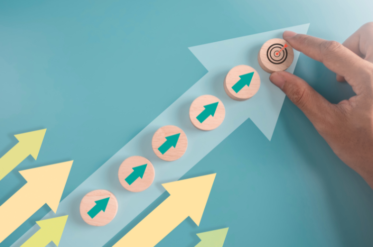 Visual representation of Succession Planning or business growth, showing five wooden discs with green arrows climbing a large blue arrow toward a final disc with a target and a hand placing the final piece.