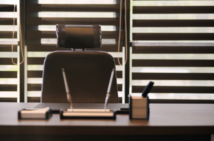 A dark, empty, adjustable executive office chair is viewed from behind, positioned at a polished wooden desk with a leather blotter, pen stand, and small desk accessories. Horizontal window blinds are visible in the background, providing dim, horizontal strips of light.