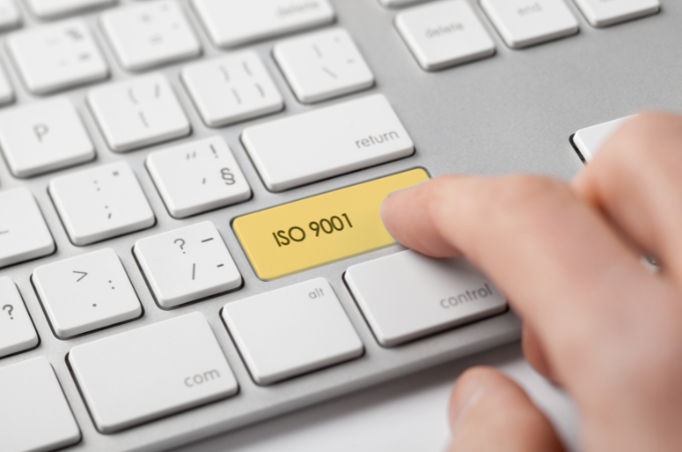 A hand presses a large, rectangular yellow key on a modern white keyboard labeled "ISO 9001", representing a focus on Quality Management Systems (QMS), compliance, or achieving certification standards.