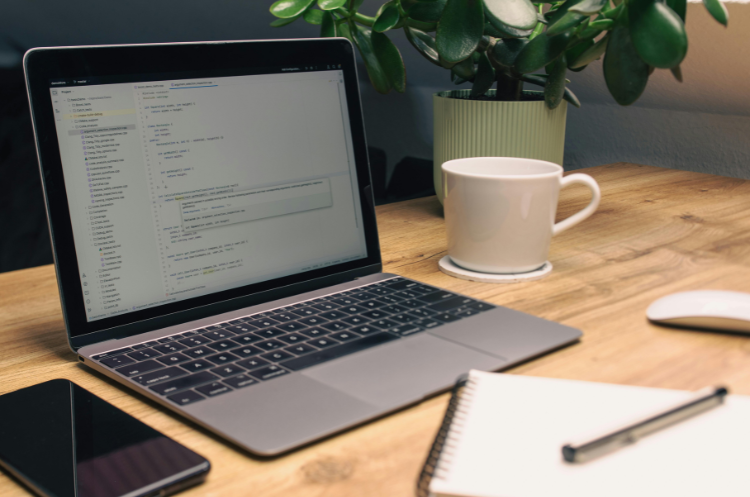 A laptop on a wooden desk displaying software code in an IDE, next to a coffee mug, notebook, and smartphone. Representing the workspace of a Chief Technology Officer (CTO) or software developer.