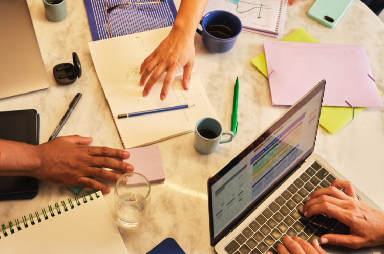 An overhead, high-angle shot of a collaborative workspace on a marble table. Three people's hands are visible, engaged in a creative or strategic planning session. One person points at a hand-drawn sketch in an open notebook, another types on a laptop displaying colorful charts, and a third rests their hand near purple sticky notes. The table is cluttered with professional tools, including a tablet, a green pen, coffee mugs, a glass of water, and various colorful folders and notebooks, creating a vibrant, productive atmosphere.