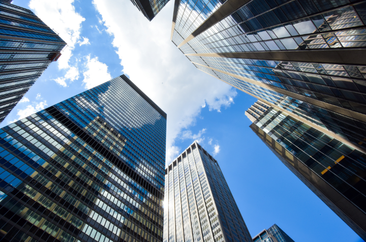 Low-angle view of modern glass skyscrapers reaching toward a blue sky with clouds. This architectural imagery represents corporate growth, business leadership, and the strategic vision of a corporate development director.