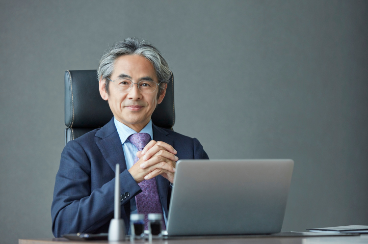 A confident, experienced male CEO in a suit sitting at an executive desk with a laptop in a Singapore office. This portrait illustrates the Chief Executive Officer role and the career transition from senior executive to CEO for leaders in Singapore.