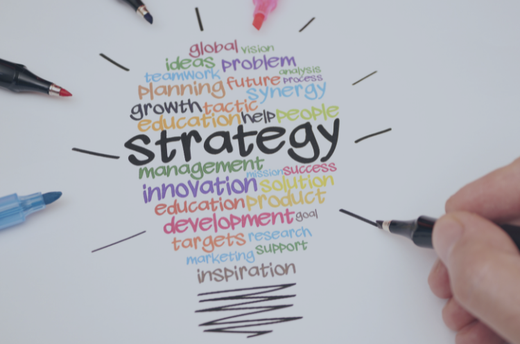 Colorful word cloud shaped like a lightbulb with “strategy” at the center, surrounded by terms like planning, innovation, growth, and development, with pens drawing around it.