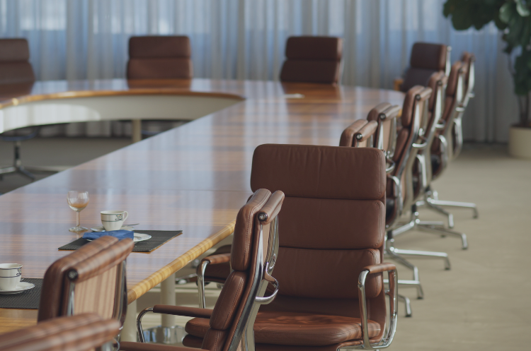 An empty, upscale corporate boardroom featuring a long wooden conference table and ergonomic leather chairs, prepared with coffee cups for a high-level meeting. The setting represents executive career transitions, board chairperson roles, and corporate governance for experienced leaders moving into senior advisory and leadership positions.