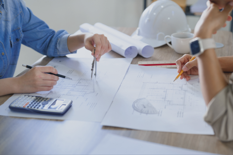 Two professionals collaborating over architectural blueprints and engineering technical drawings on a wooden desk. The scene includes tools such as a compass, calculator, and a white hard hat, highlighting the transition from technical field work to engineering consulting, project management, and strategic infrastructure planning.