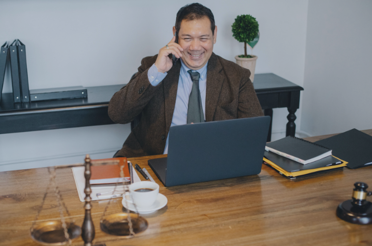 A smiling, experienced consultant in Singapore sitting at a wooden desk, engaged in a professional phone call while working on a laptop. The office setting includes legal scales, a gavel, and professional documents, representing expert advisory services, legal consulting, and career opportunities for senior professionals in the Singapore job market.