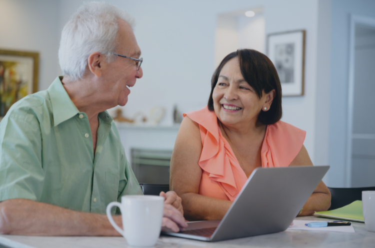 Two experienced professionals in Singapore smiling and collaborating while using a laptop in a relaxed home office setting. This image represents the benefits of flexible work opportunities, remote consulting, and work-life balance, highlighting how senior talent can leverage digital tools to find part-time or project-based roles in the Singaporean job market.