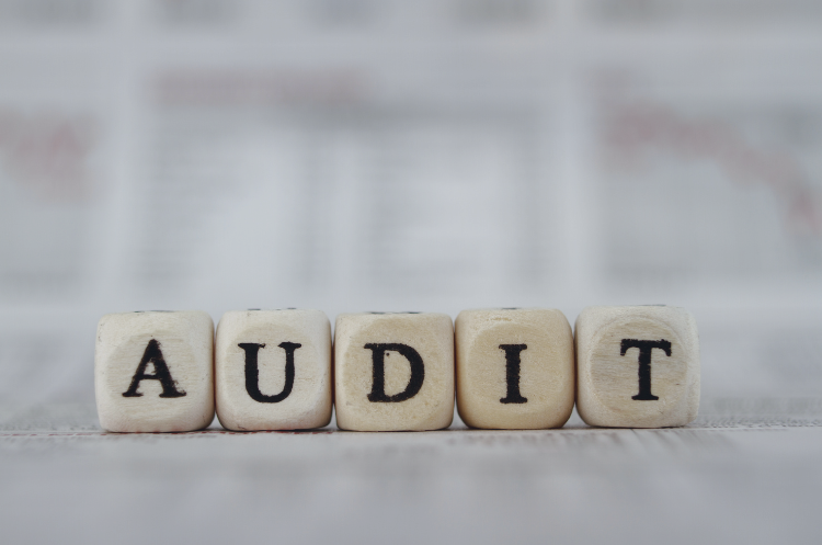 Wooden letter blocks spelling "AUDIT" placed on a desk with blurred financial reports in the background, representing internal audit, governance, compliance, and risk management.