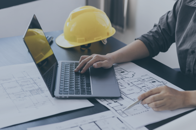 Engineer reviewing building plans and working on a laptop with a safety helmet on the desk, illustrating engineering jobs in Singapore