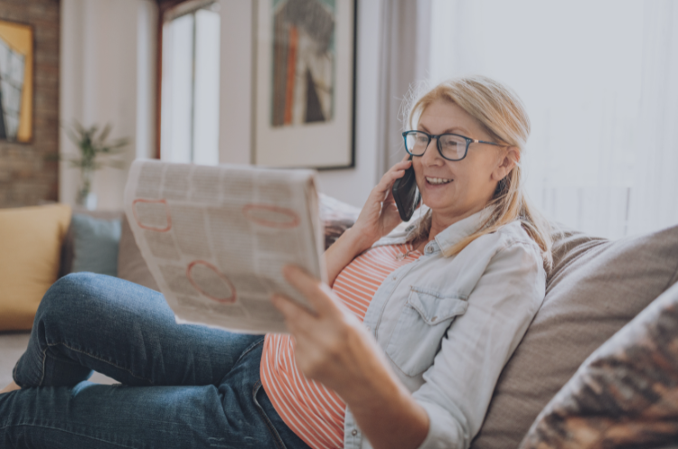 Senior woman reading a newspaper while talking on the phone at home, representing flexible part-time jobs for seniors in Singapore.