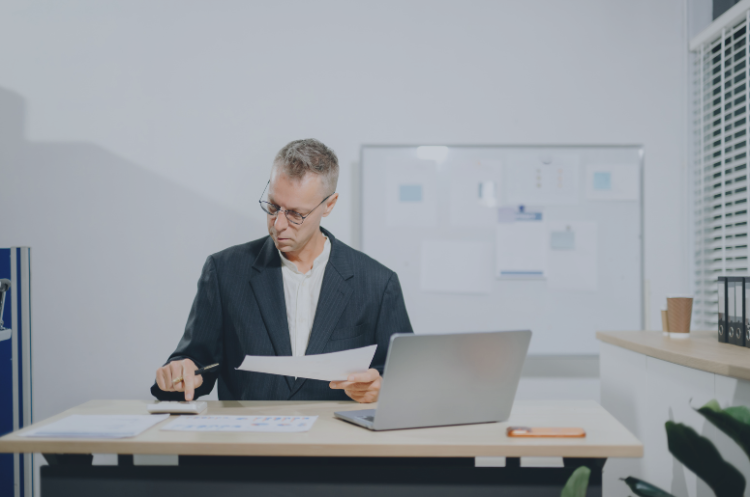 Experienced professional reviewing documents at a desk with a laptop in an office.