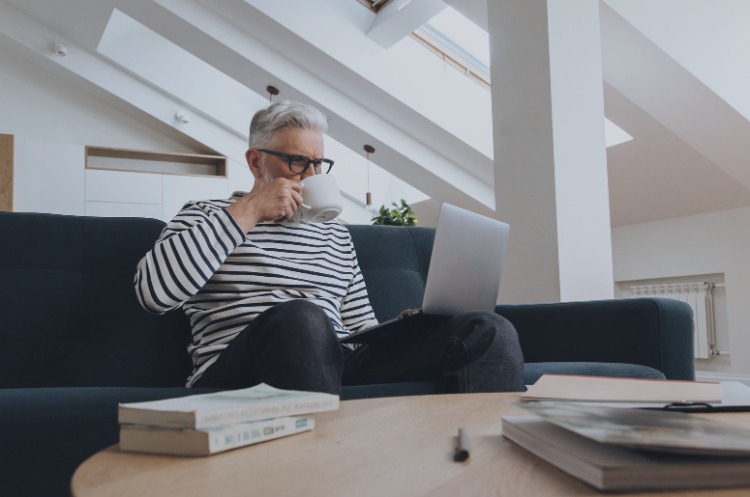 A gray-haired man wearing glasses sits on a sofa, drinking from a mug while using a laptop in a bright, modern living room. A coffee table with books and a pen is in the foreground.
