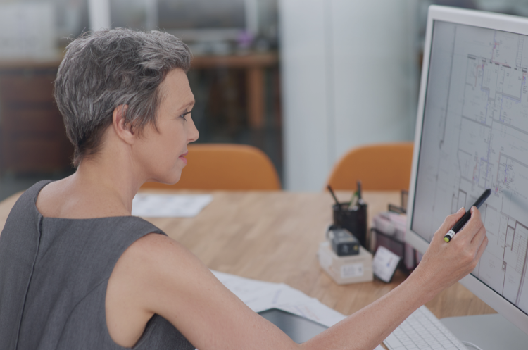 A woman working at a computer, reviewing architectural plans displayed on a large monitor.