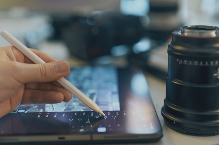 A hand using a stylus on a digital tablet beside a camera lens, representing digital technology and business transformation.